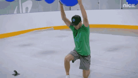 a man in a green shirt is throwing a blue ball on a rink with nick written on the wall behind him