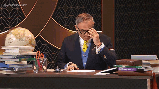 a man in a suit sits at a desk with a stack of books and a globe behind him