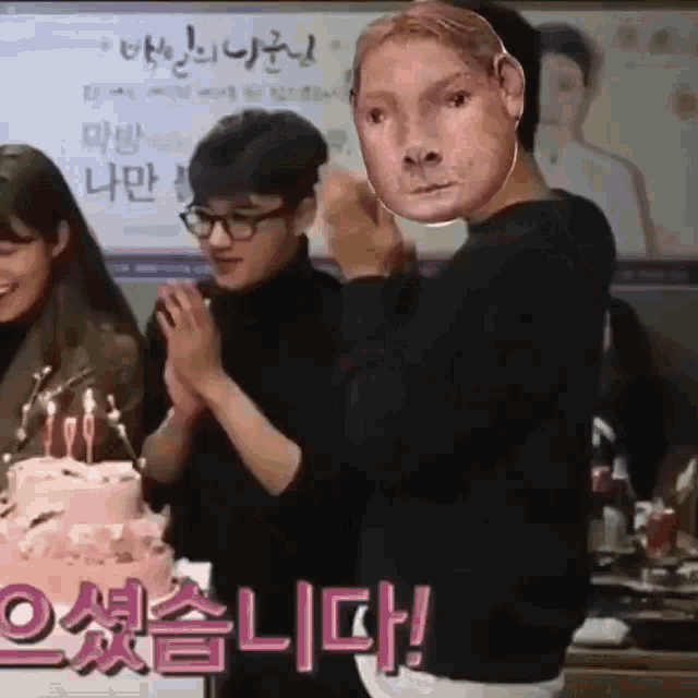a group of people are standing in front of a cake with candles and a man with a mask on his face