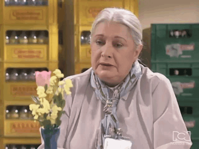 an elderly woman is holding a vase of flowers in front of a bunch of crates of beer .