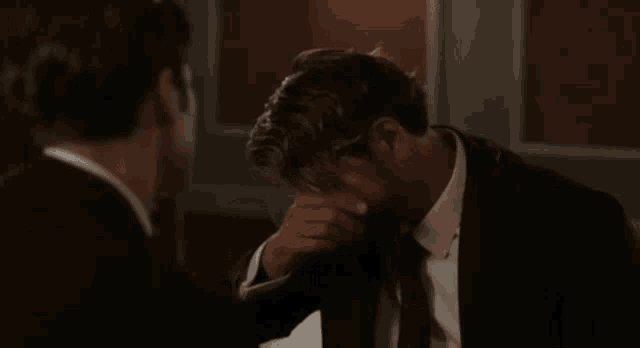a man in a suit and tie is touching another man 's head with his hand .