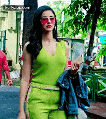 a woman in a green jumpsuit and pink sunglasses is walking down a street .
