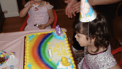 a little girl wearing a party hat looks at a birthday cake