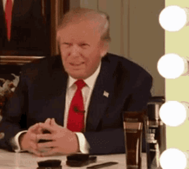 donald trump is sitting at a table with his hands folded in front of a mirror and looking at his phone .