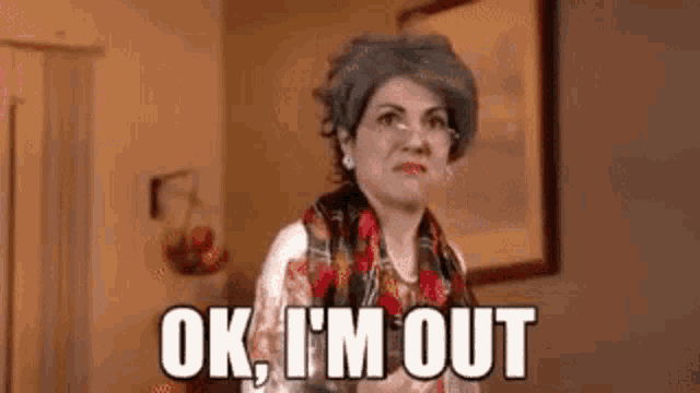 a woman in a wig and scarf is standing in a living room and says `` ok , i 'm out ''