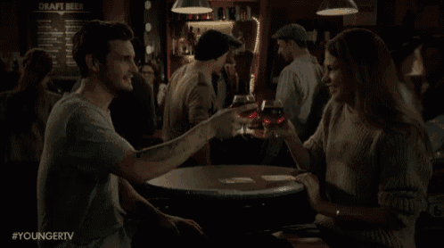 a man drinking a beer next to a woman sitting at a table with a sign that says draft beer