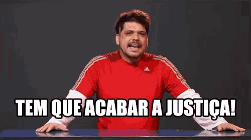a man in a red shirt is standing in front of a table with the words tem que acabar a justica written on it .