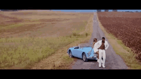 a man and a woman are standing next to a blue sports car on a dirt road .