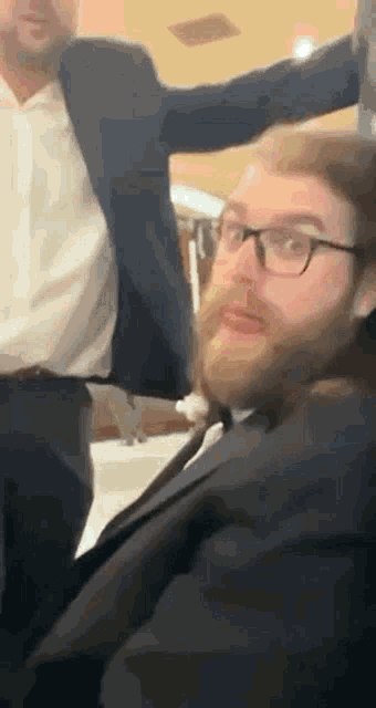 a man with a beard wearing glasses and a suit is sitting next to another man .