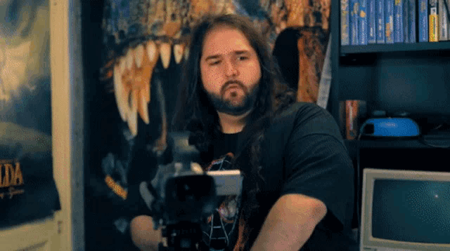 a man with long hair and a beard stands in front of a wall with a poster that says zelda