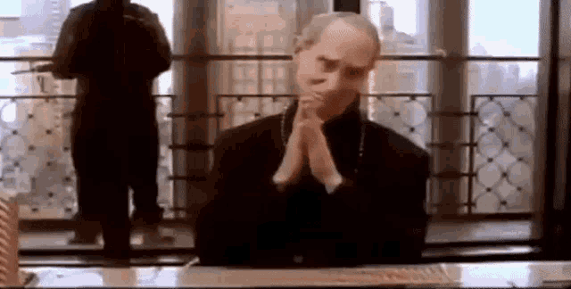 a man is sitting at a desk with his hands folded in front of a window .