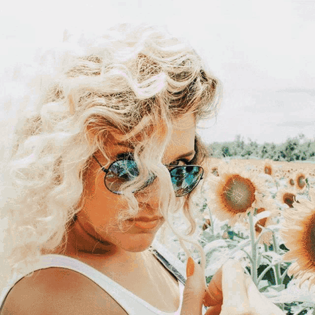a woman with curly blonde hair wearing sunglasses stands in a field of sunflowers