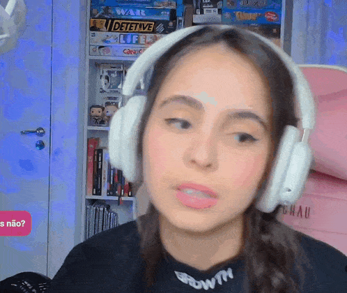 a woman wearing headphones looks at the camera in front of a shelf full of board games including detective life and war