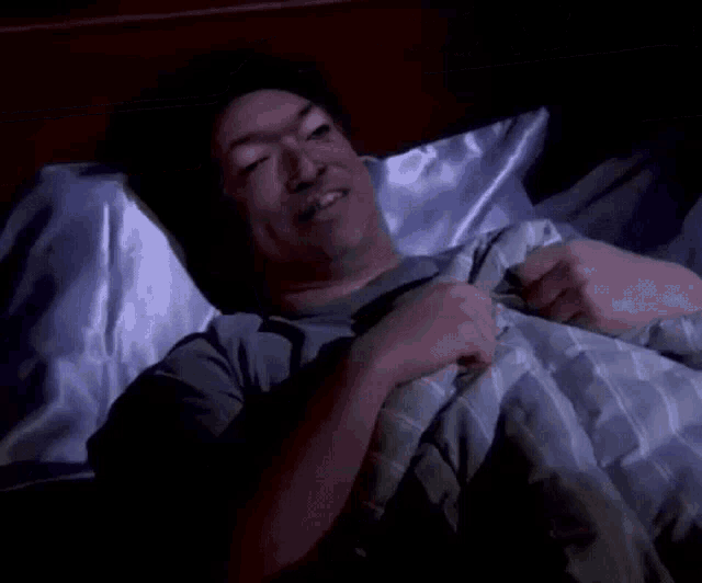 a man is laying in bed with a pillow and a blanket and smiling .