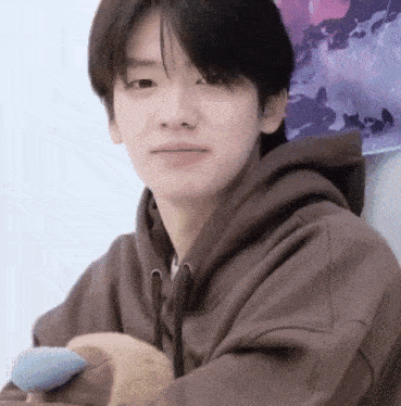 a young man wearing a brown hoodie is holding a stuffed animal and looking at the camera .
