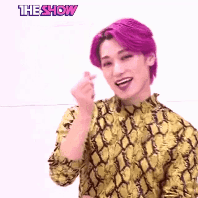 a young man with purple hair is wearing a yellow snakeskin shirt and making a heart shape with his hands .
