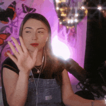 a woman wearing overalls and ear buds waves her hand
