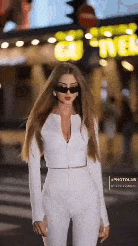 a woman wearing a white jumpsuit and sunglasses is standing on a street .
