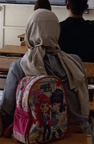 a girl wearing a hijab sits in a classroom with a backpack that says strawberry shortcake on it