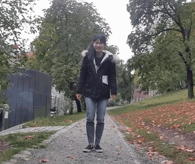 a woman wearing a black jacket and jeans is walking down a path in a park