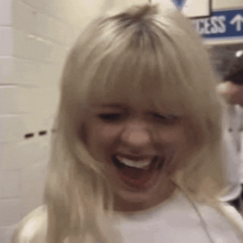 a woman with blonde hair is laughing with her mouth open in front of a blue sign that says access
