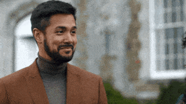 a man with a beard wearing a brown suit and a grey turtleneck