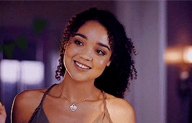 a woman with curly hair is smiling and wearing a necklace with a heart pendant