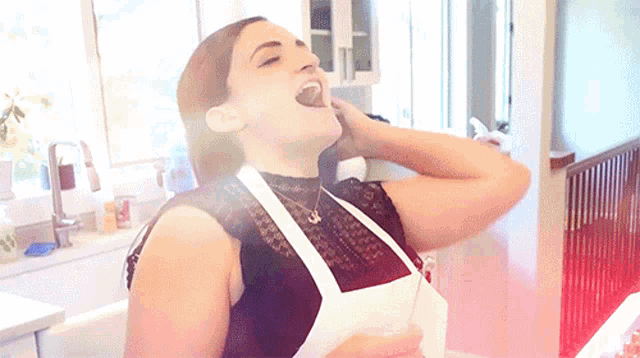 a woman in a black top and white apron is eating something in a kitchen
