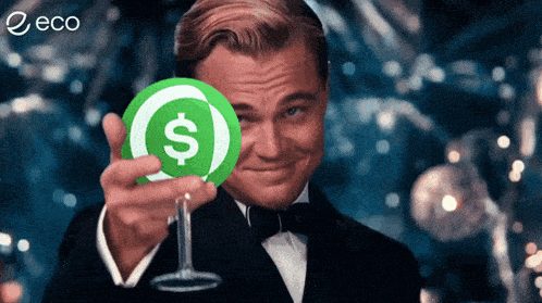 a man in a tuxedo holds a green coin with a dollar sign on it in front of his face