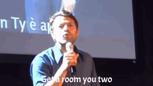 a man speaking into a microphone with the words " get a room you two " behind him