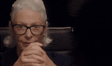 an older woman wearing glasses and earrings sits with her hands folded in front of her face