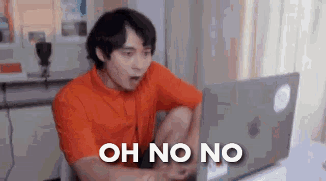 a man in an orange shirt is sitting in front of a laptop computer and saying `` oh no no '' .