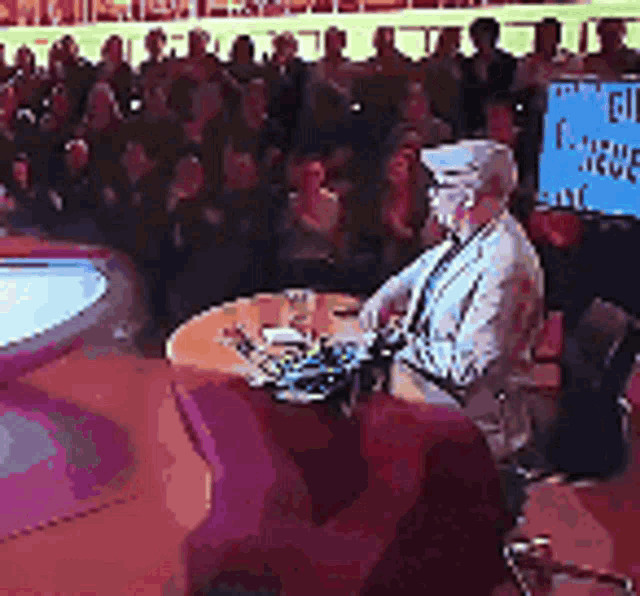a man is sitting at a table in front of a crowd with a sign that says ' i love you '