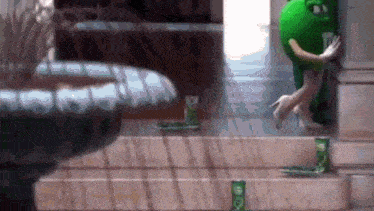 a green m & m 's mascot is standing on a set of stairs next to a fountain