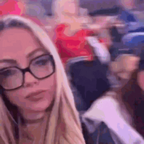 a blonde woman wearing glasses is sitting in a crowded room