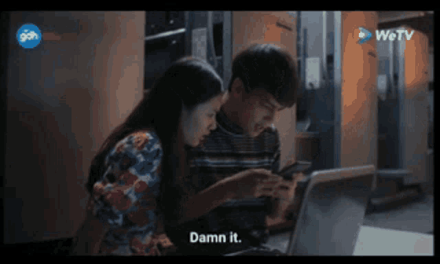 a man and a woman are sitting in front of a laptop and the woman says damn it