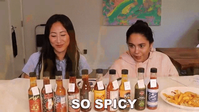 two women are sitting at a table with bottles of hot sauce and the words so spicy written on the table