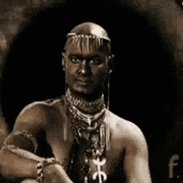 a man without a shirt is sitting in a dark room wearing a necklace .