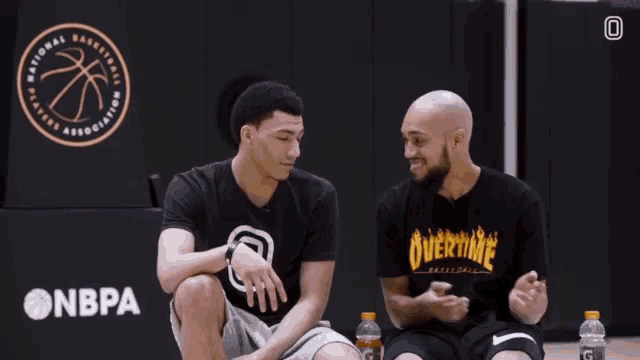 two men are sitting on a basketball court and one has a shirt that says overtime on it