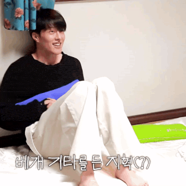 a man in a black sweater sits on a bed with korean writing on the bottom right corner