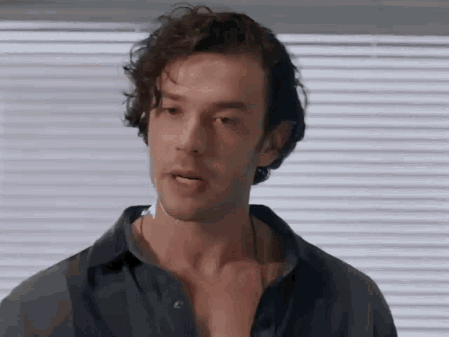 a man with curly hair is standing in front of a window with blinds and making a funny face .