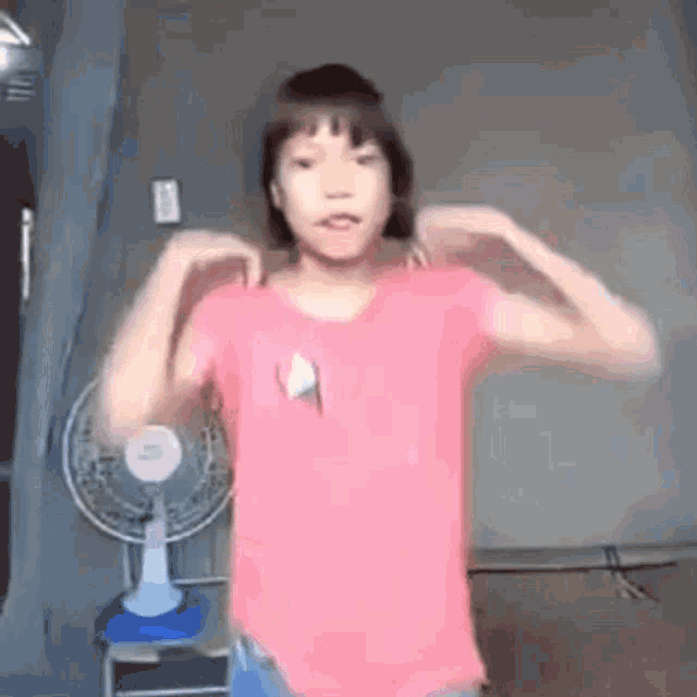 a young girl in a pink shirt is standing in front of a fan and flexing her muscles .