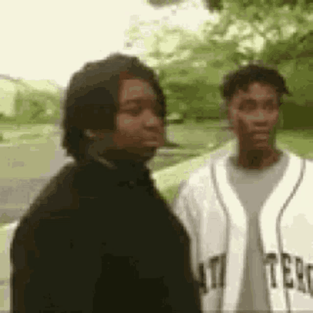 two men are standing next to each other in a park . one of the men is wearing a baseball jersey .