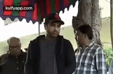 a man in a baseball cap is talking to another man in a plaid shirt while standing next to a tree .