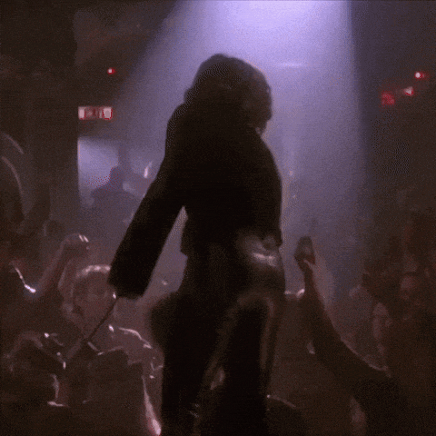 a woman is dancing in front of a crowd in a dark room with a red exit sign in the background