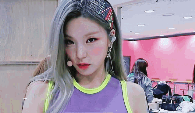 a woman wearing a purple tank top and earrings is standing in front of a pink wall .