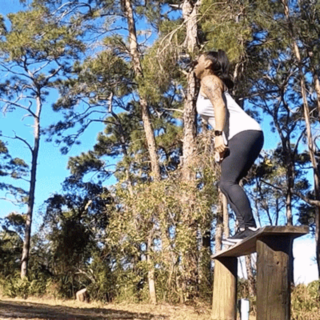 a woman in a white tank top and black leggings is squatting on a wooden post