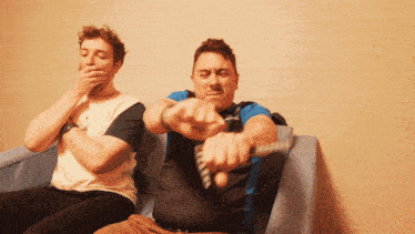 two men sitting on a couch one covering his mouth and one holding a remote control