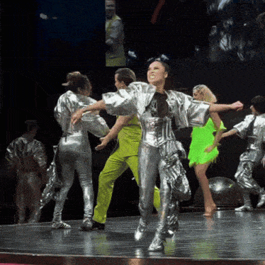 a group of people are dancing on a stage and one of them is wearing a silver outfit