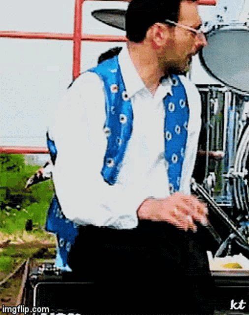a man wearing a blue vest and a white shirt playing drums